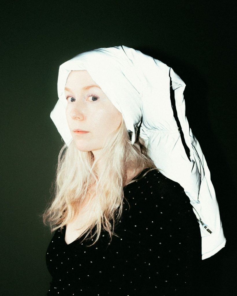 A caucasian woman with blonde hair, wearing a white headscarf, in black top, posing against a dark backdrop