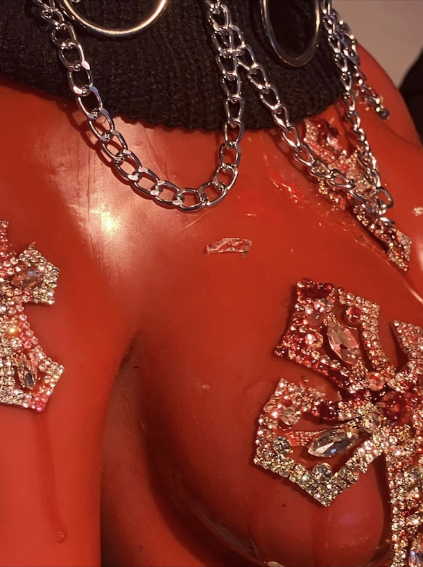 a close up of the plastic body of a pink sex doll that has been covered in chains and bejewelled cross shapes. There is also a stray shard of glass attached to the "body" of the doll