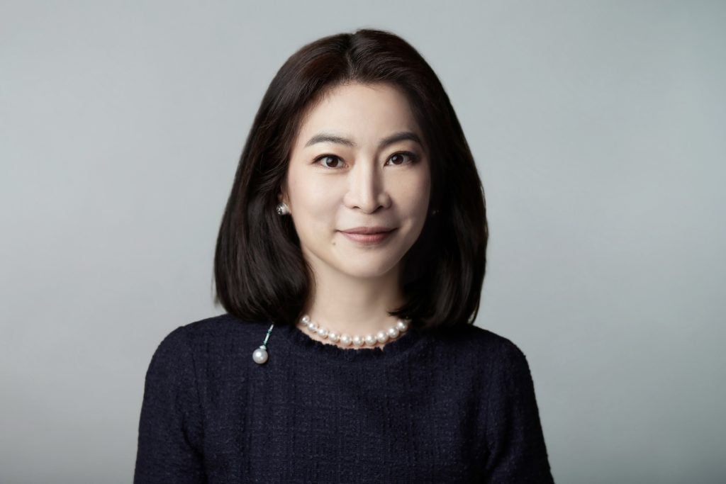 An East Asian woman with shoulder length dark hair, in a navy top, pearl necklace, posing against a grey backdrop.