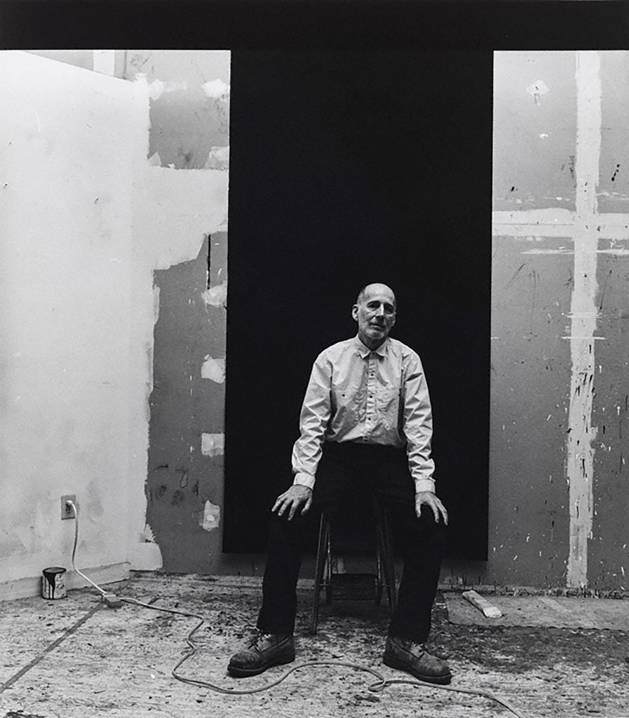 A black and white photo of a young Robert Moskowitz in his Tribeca Studio, New York. He is sitting is a raw looking space with large paintings leaning against the walls. 