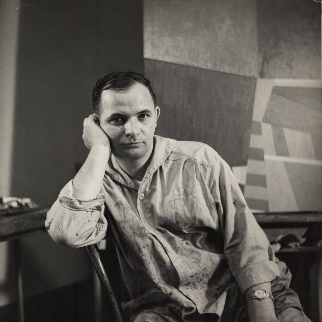 A photo of a young white man with short dark hair in a paint splattered collared shirt, leaning his chin on his right hand. There are large paintings resting against the wall behind him.. 