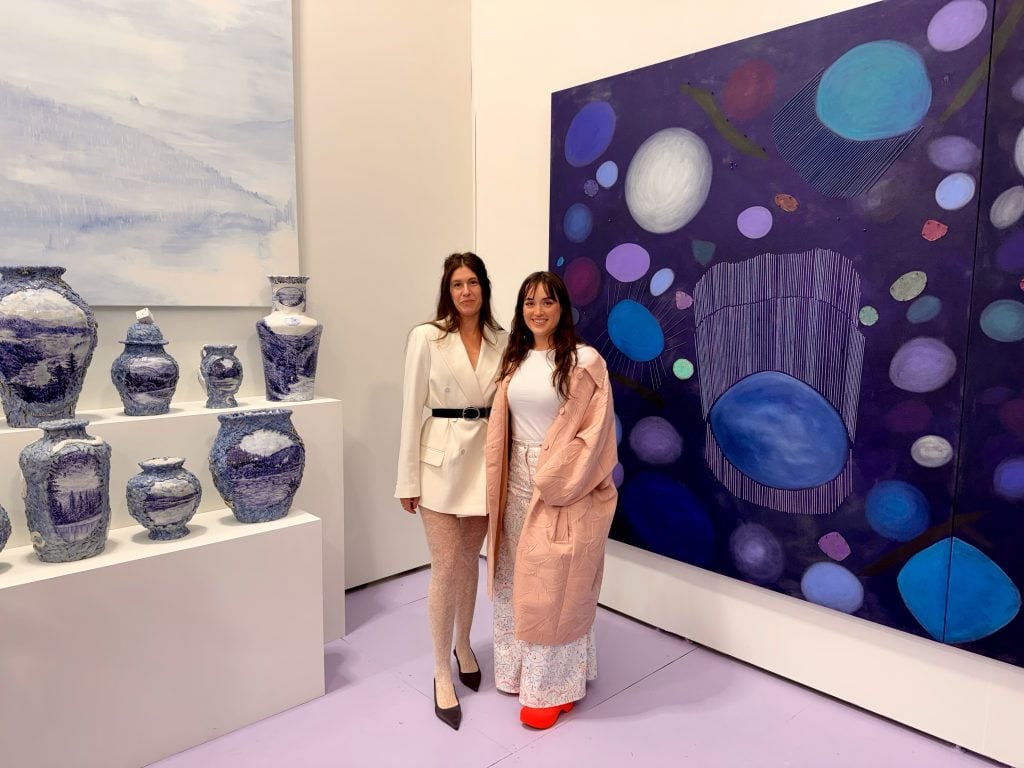 Sachi Moskowitz and Lacey Stoffer Anna Erickson Presents. They are two young women with dark hair, standing between a stand of blue and white vases and a large purple and blue abstract canvas. 