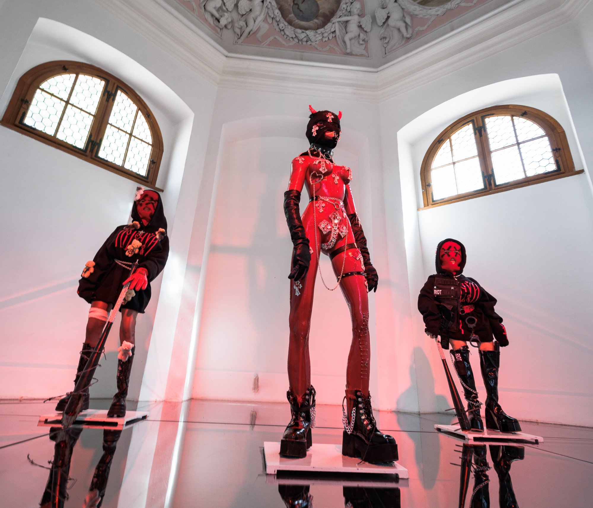 a white-walled interior space of a small chapel filled with three female dolls wearing pink and black outfits installed standing in a v formation