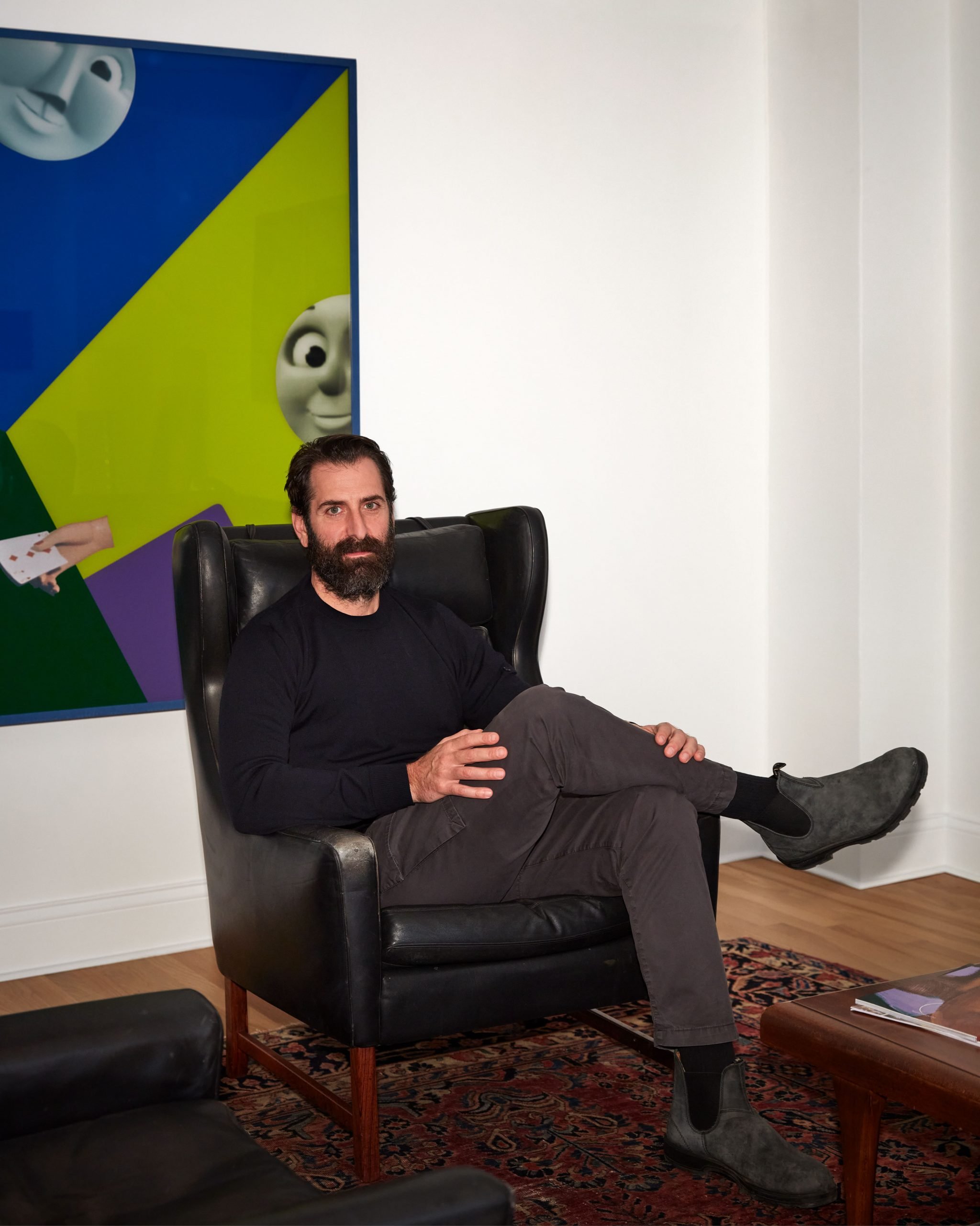 A man, art advisor Joe Sheftel, sits in a modern chair in a white room in front of a painting crossing his legs