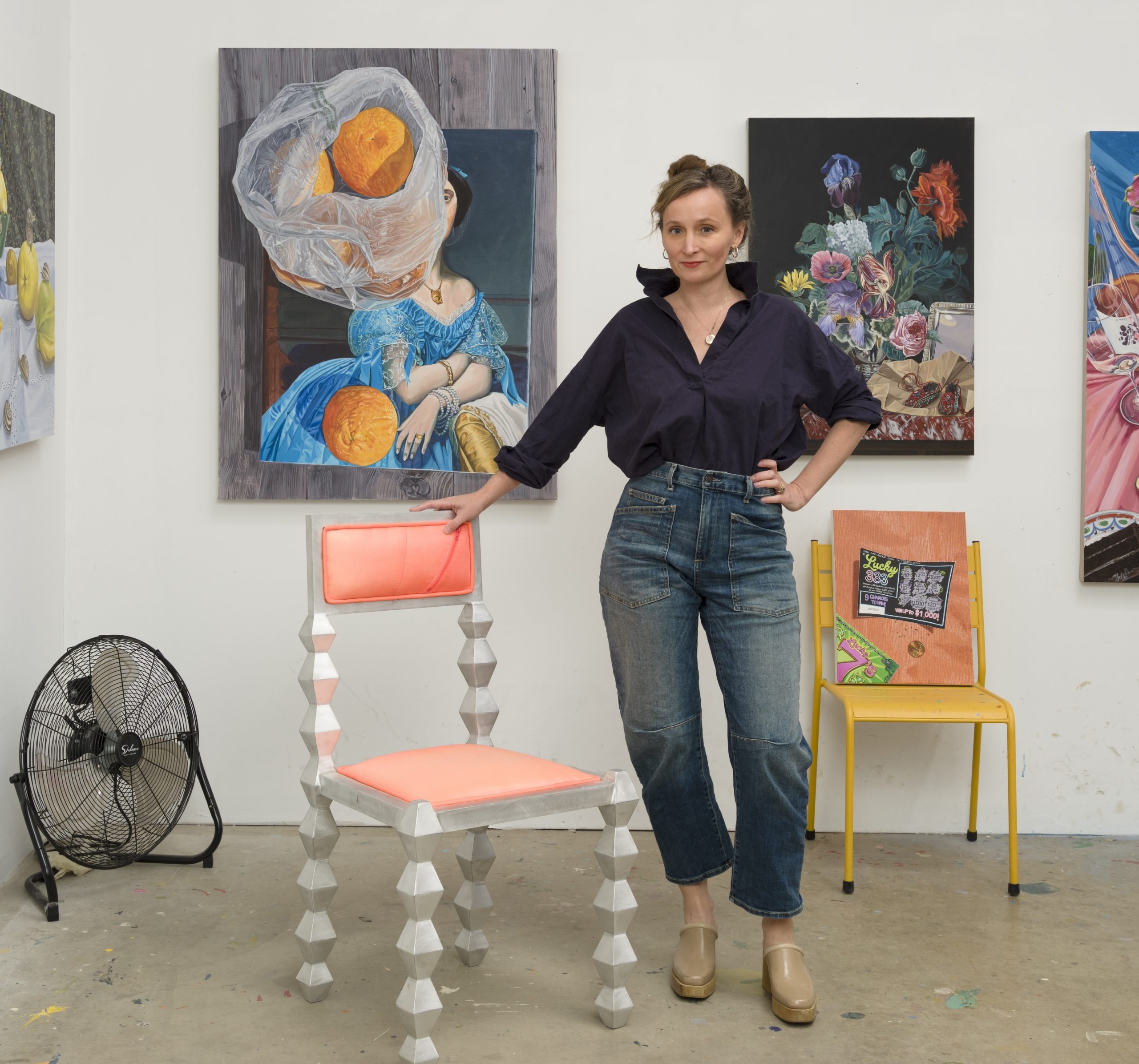 a white woman in jeans and a black blouse stands in her studio in front of paintings of still lifes