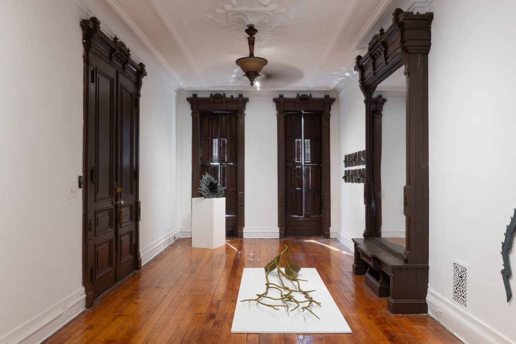 "Helen Evans Ramsaran: Strange Fruit," installation view. A bronze statue of a fallen branch lying on a low platform on the wooden floor of a 19th-century drawing room with a large mirror in an ornate, floor-to-ceiling mirror. There are matching wooden frames on the floor to ceiling windows, and the door to the entry opposite. The walls and white and the ceiling has some decorative molding. 