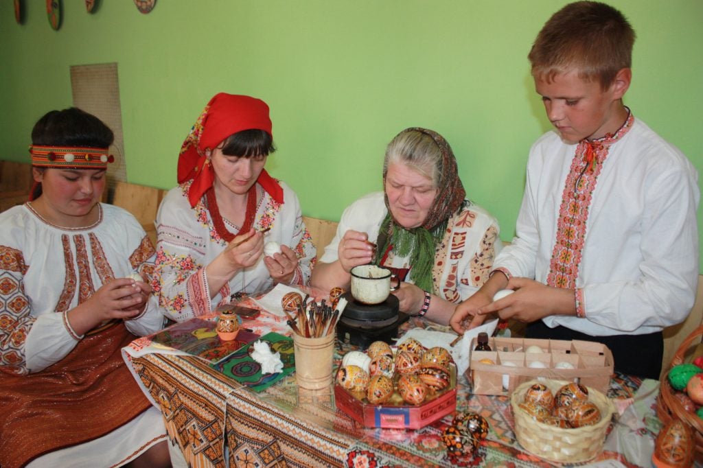 a multigenerational group of people in traditional dress sit in an interior space at a wooden table painting eggs, there are painted eggs all over the table and supplies for painting