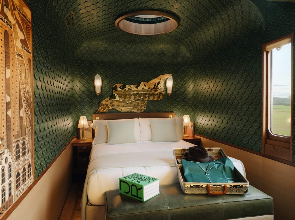 an epaulet green and gilded train car features a bed and a green leather trunk has an open old fashioned suitcase atop it 