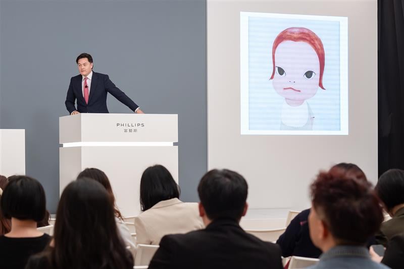 a man stands at an auction podium with a large painting of a child behind him