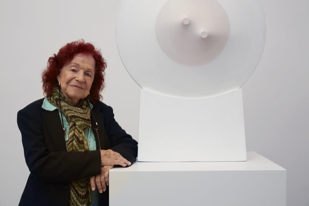 Zilia Sánchez with her work at Galerie Lelong & Co., New York, 2014. She is an elderly woman with reddish hair, standing next to a circular sculptural white and baby pink canvas work that protrudes from the wall.