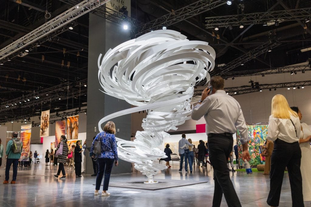 A large, swirling white sculpture is the centerpiece at an art exhibition, surrounded by attendees admiring various artworks in a spacious venue.