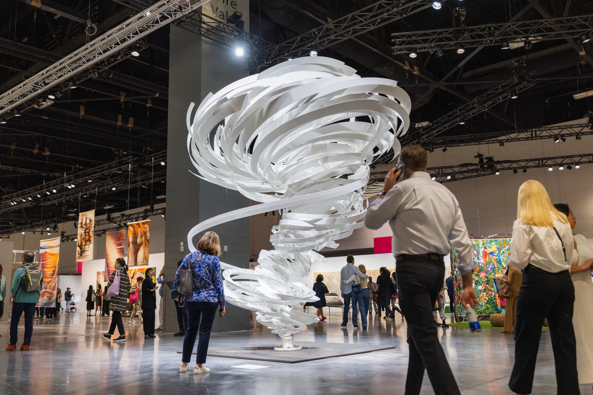 A large, swirling white sculpture is the centerpiece at an art exhibition, surrounded by attendees admiring various artworks in a spacious venue.