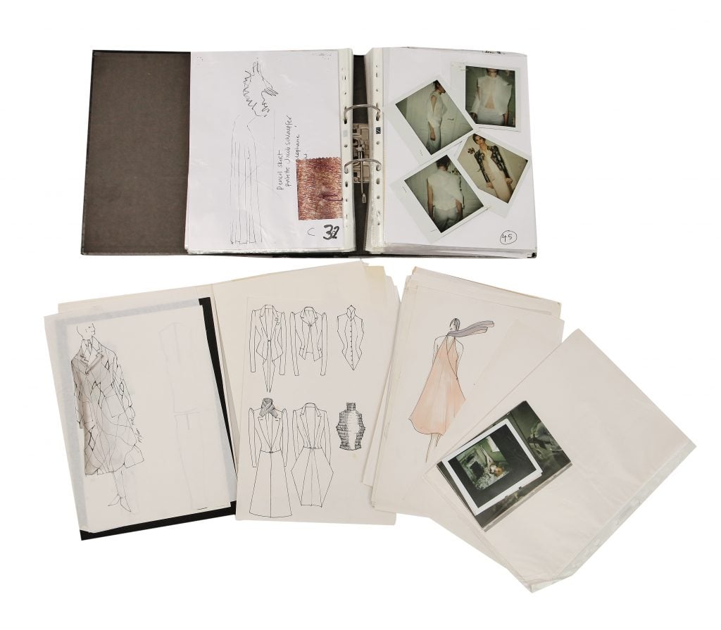 An aerial view of an open sketchbook with Polaroid photos inside of it, as well as additional pages with fashion illustrations below it, all on a plain white background.