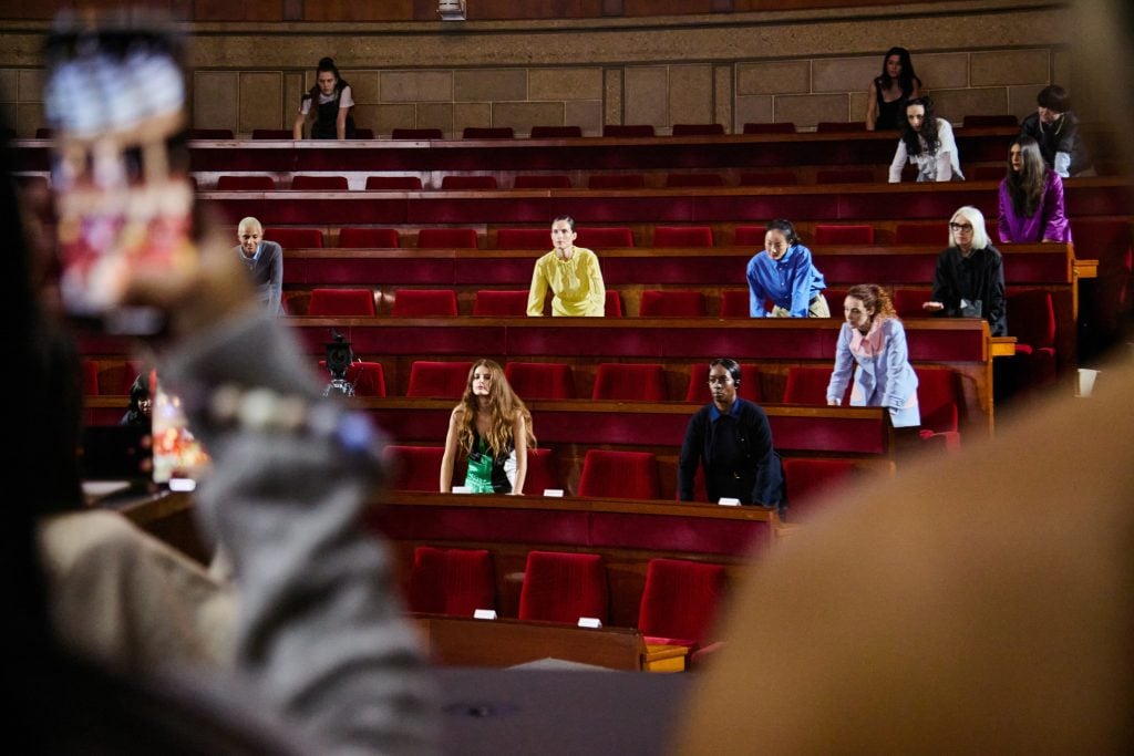 Performers in vibrant attire seated and standing in a tiered theater with red velvet seats.