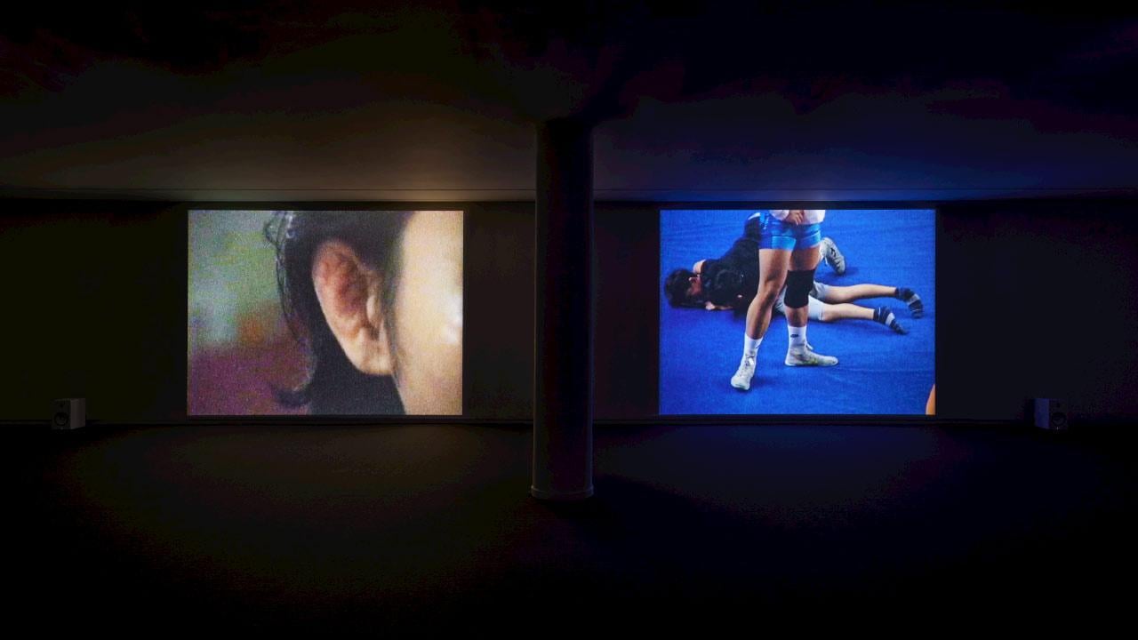 Two screens in a dark room show bodies—one a closeup of a face, the other of two wrestlers