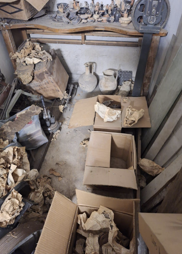 A photograph depicting scores of stolen Ancient Greek relics like vessels scattered about throughout surfaces and brown cardboard boxes in a basement.