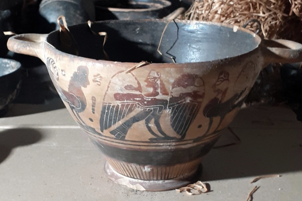 A photograph of a brown Ancient Greek vessel engraved with black and reddish designs of human-birdlike figures.