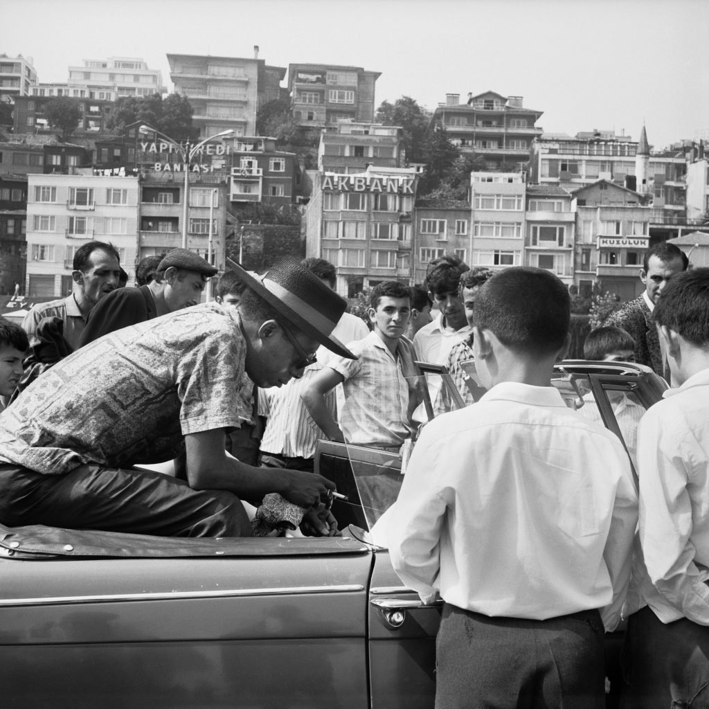 black and white photograph of a black man in a car with a hat interacting with a group of people wearing white shirts with a nice view of the city in the background