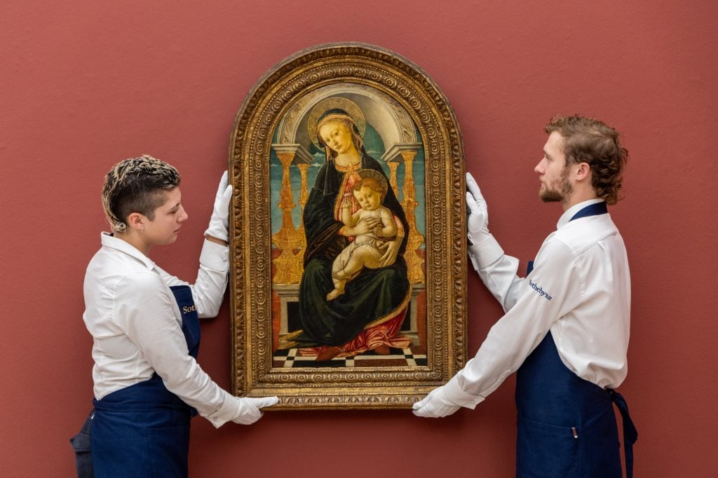 Two people dressed in white shirts and black trousers holding a painting in a gold frame up from either side in front of an earthy red wall.