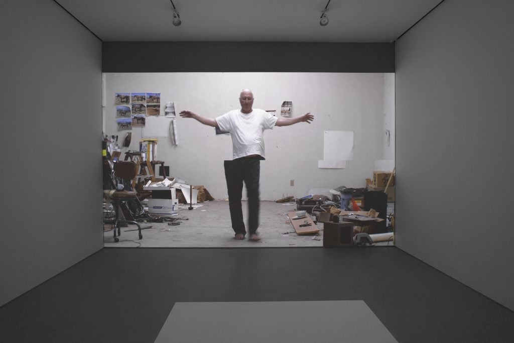 A darkened art gallery with a video of the artist Bruce Nauman standing in his studio, arms outstretched