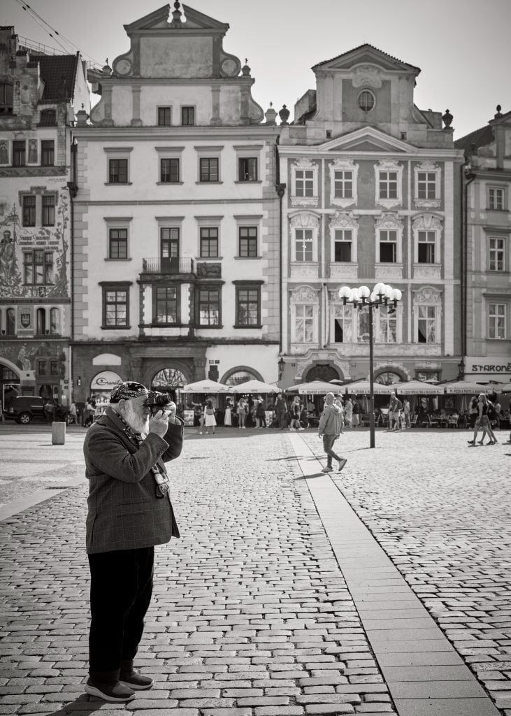 A man with a camera stands on a cobblestone street in a historic European square.