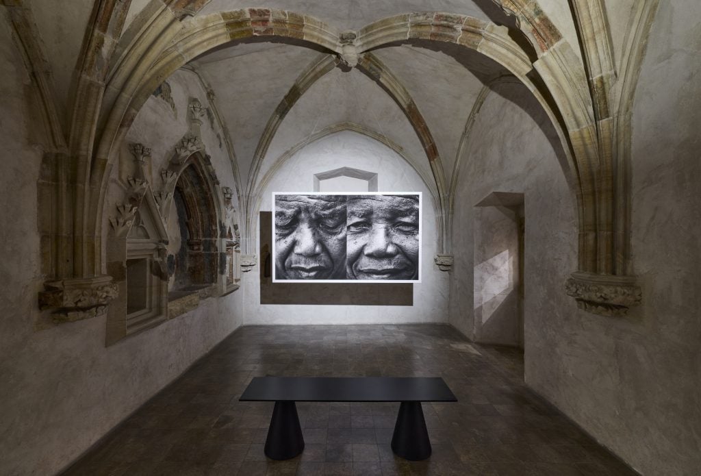 A black-and-white photo installation of an elderly face displayed in a gothic stone gallery.