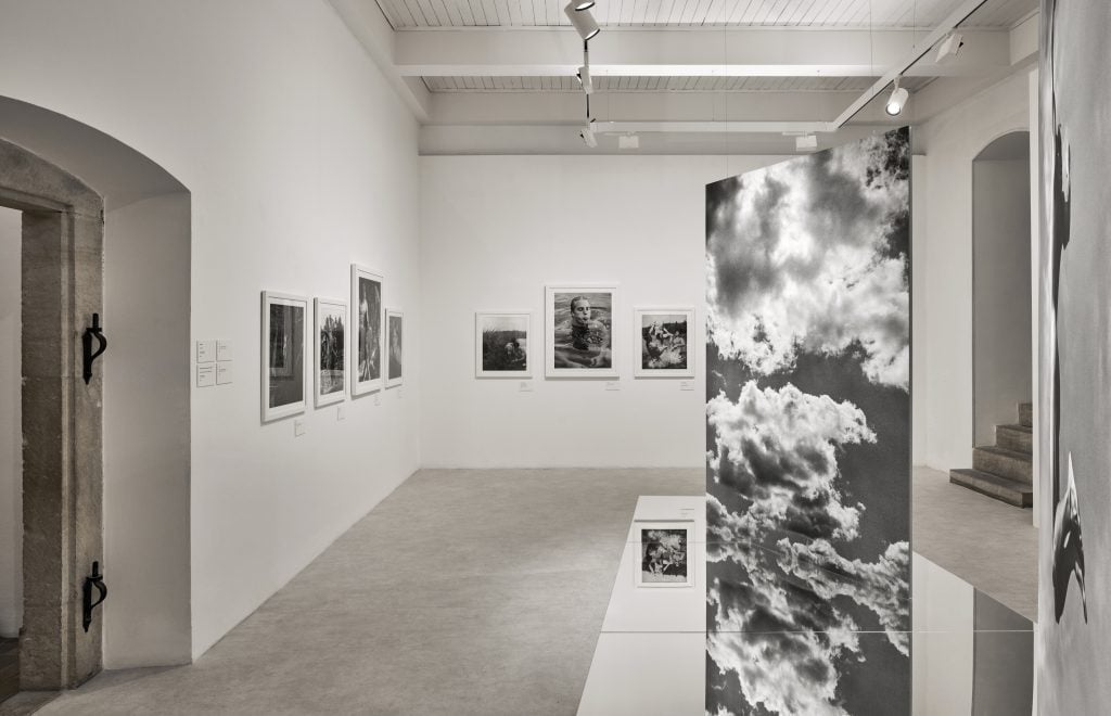 A contemporary exhibition room with striking black-and-white photographs and a reflective panel featuring cloudy skies.