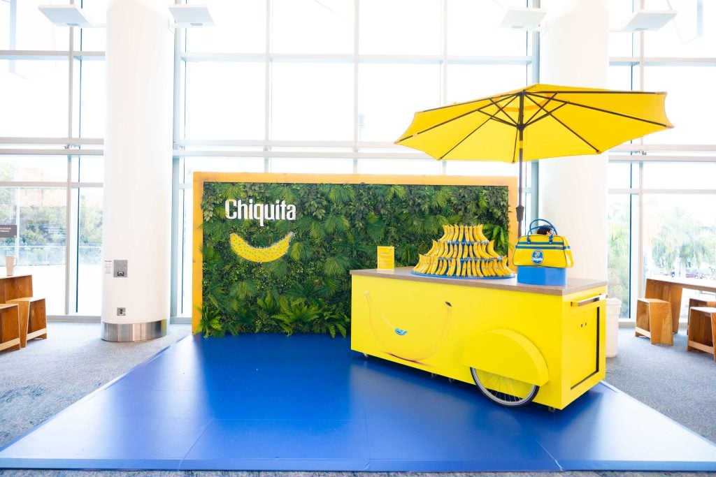 The Chiquita banana stand at Art Basel. There is a blue floor, a yellow cart with a yellow umbrella and a display of free bananas, and a leafy green wall.