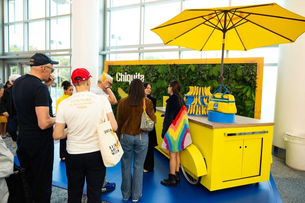 Crowds at the Chiquita banana stand at Art Basel. There is a blue floor, a yellow cart with a yellow umbrella and a display of free bananas, and a leafy green wall. 