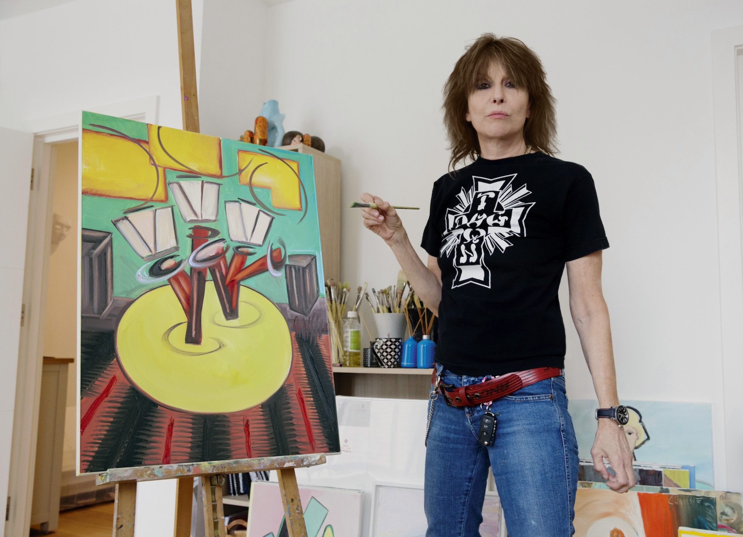 Musician Chrissie Hynde stands in front of an easel with a painting, paintbrush in hand