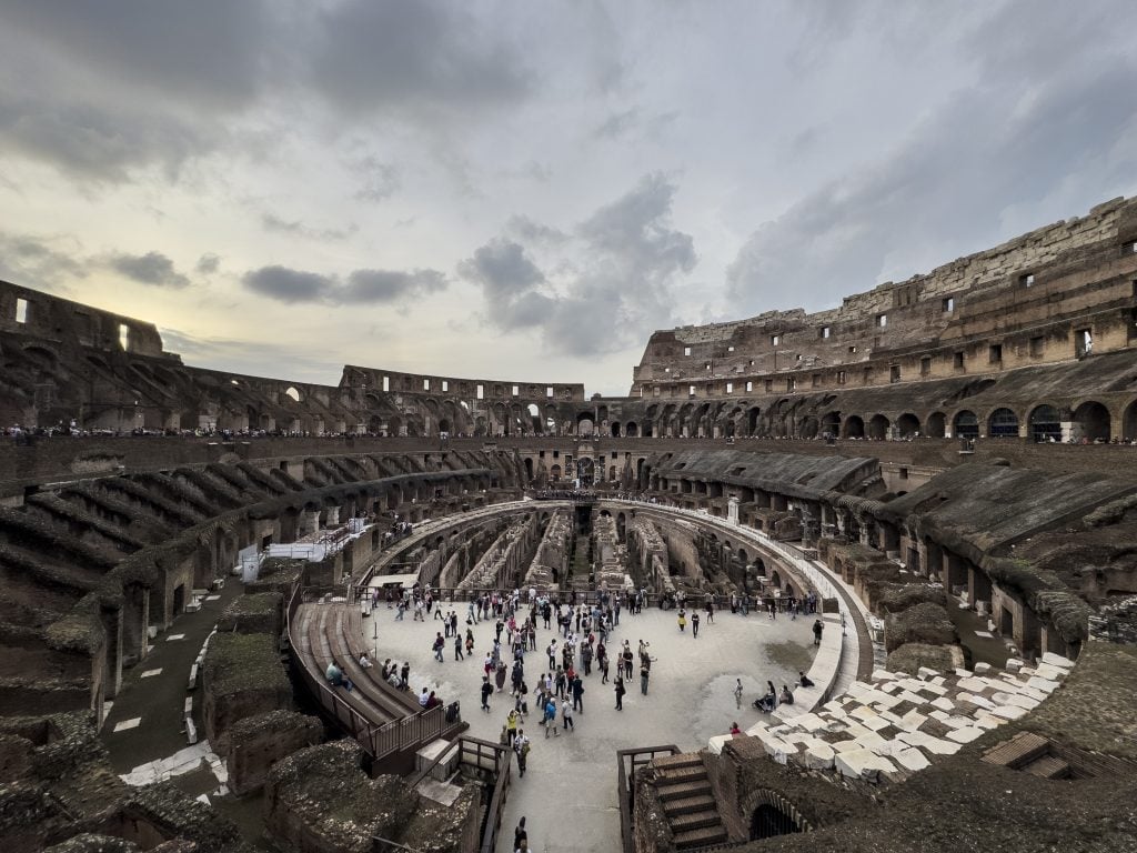 photograph of the collosseum in rome showing a large round structure with various dillapidated arches and underground pathways in which tourists from all corners of the world can admire the beauty of ancient rome