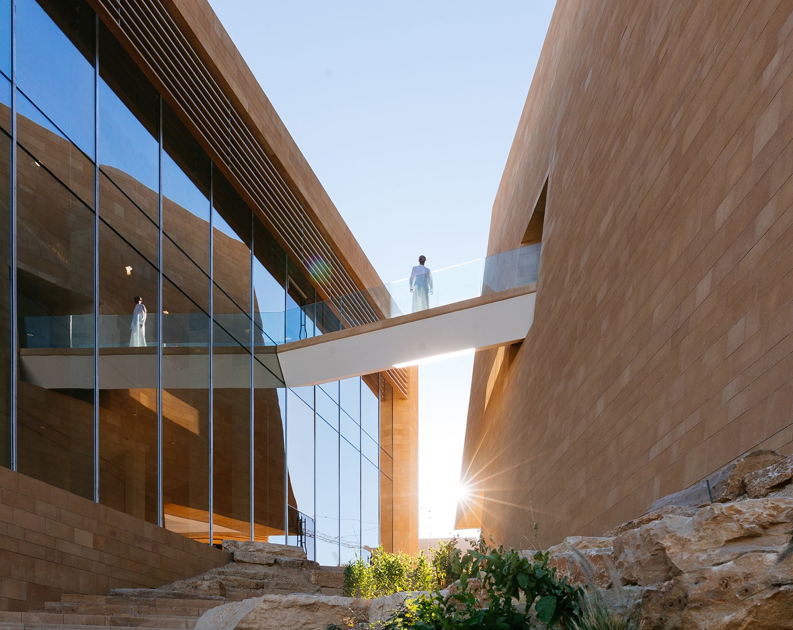 A modern architectural space featuring angular sandstone walls, glass reflections, and a suspended walkway with individuals, illuminated by natural light and framed by rocky landscaping, home to Diriyah Art Futures.