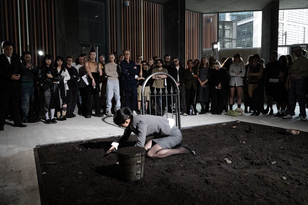 A crouched performer digs into dirt within an installation, as an audience attentively observes.