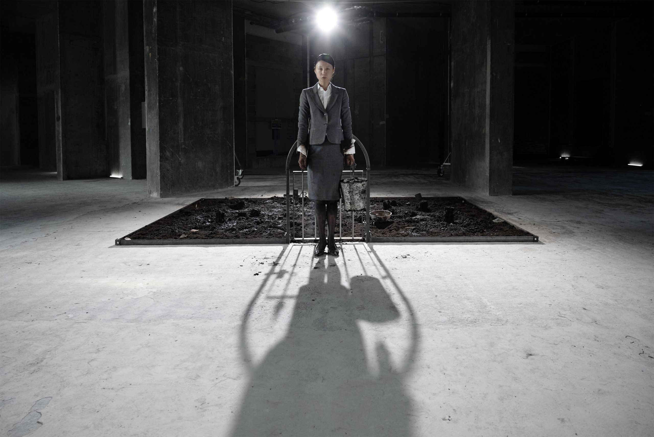 A performer in a suit stands solemnly before a shadowed dirt-filled installation under spotlight.