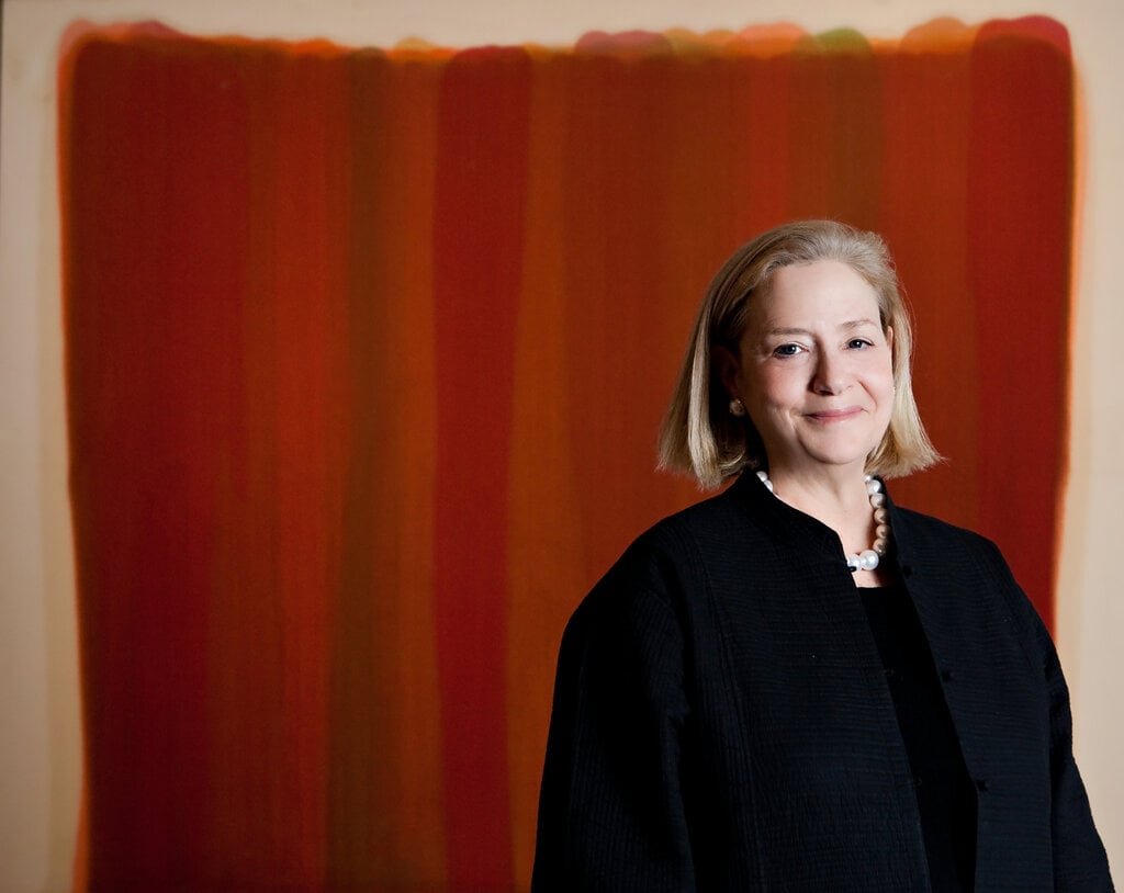 A photo of Hope Alswang, a middle-aged white lady with shoulder length blonde hair, wearing a large black shirt and white beaded necklace, stands in front of an abstract red color field painting. 