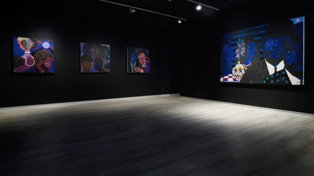 Installation view of "Stan Burnside: Blacks and the Blues" (2024–25) at Mestre Projects in Nassau showing four paintings installed on black gallery walls and spotlit.