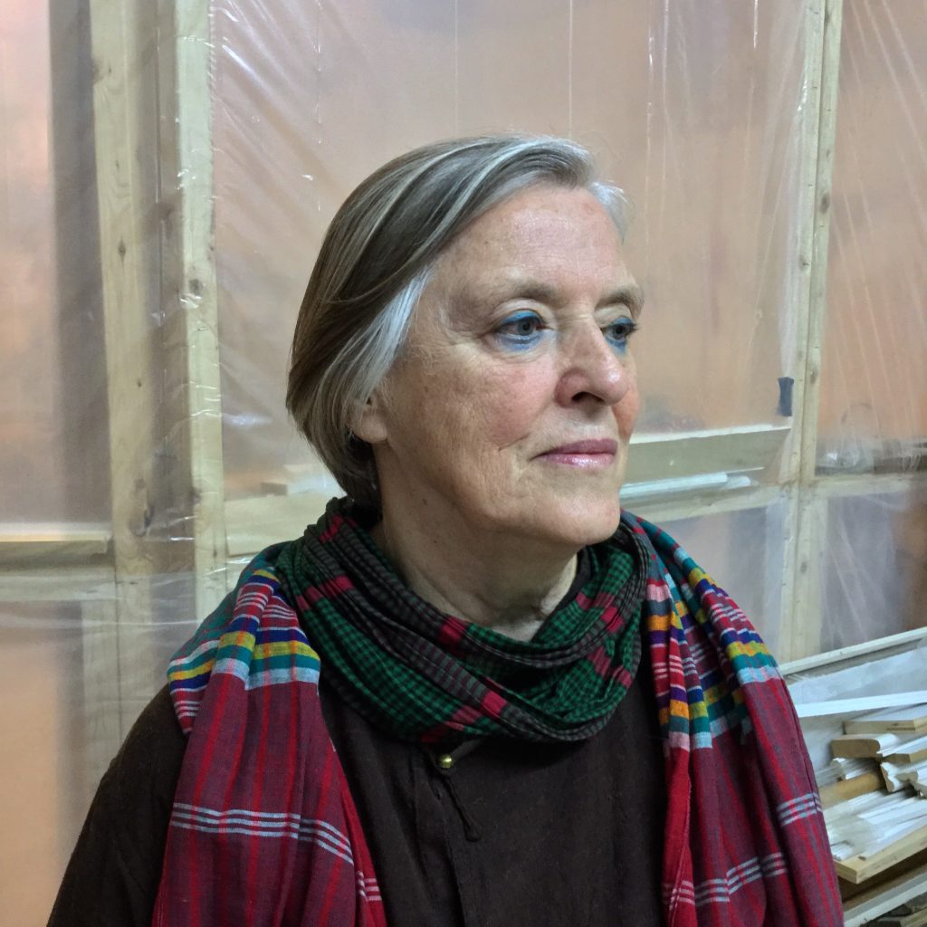 Jackie Windsor in 2019. A color photo of an elderly woman with short gray hair wearing a red plaid scarf, seen from the mid chest up in front of the backs of several painting canvases.