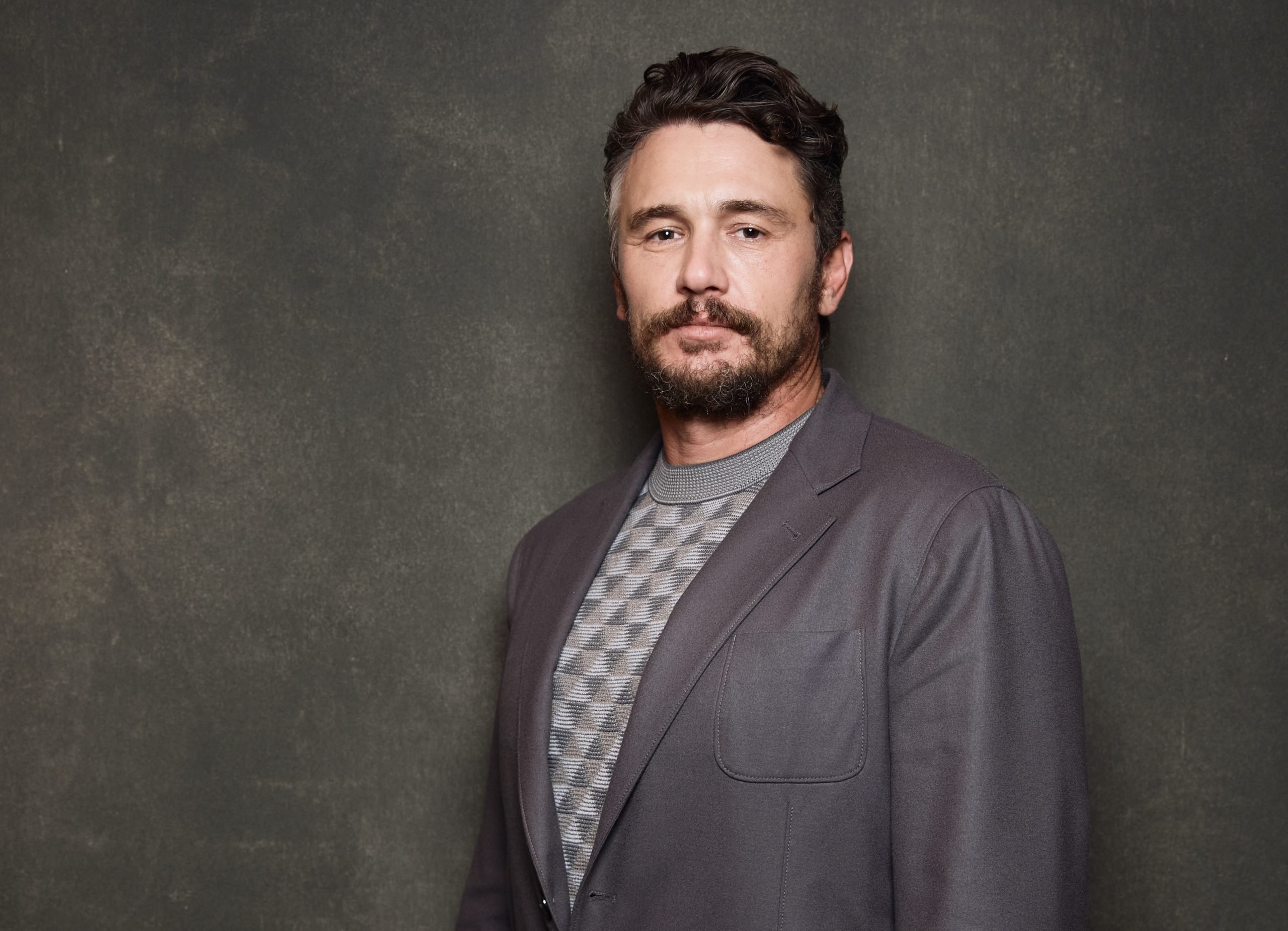 Actor James Franco posing in a patterned grey sweater with grey blazer against a dark backdrop