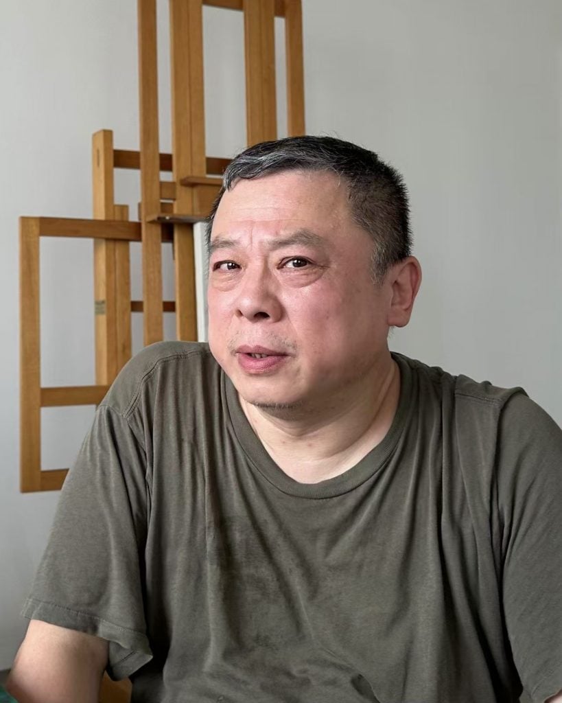 Photo of artist Ji Dachun in an O.D. green teeshirt sitting in front of an empty easel in a white studio space.