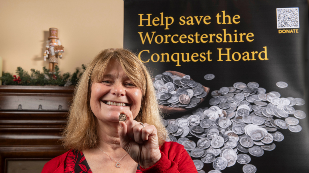 A. photograph of a middle aged blonde woman holding a silver coin up, with a sign that says 'Help save the Worcestershire Conquest Hoard' behind her