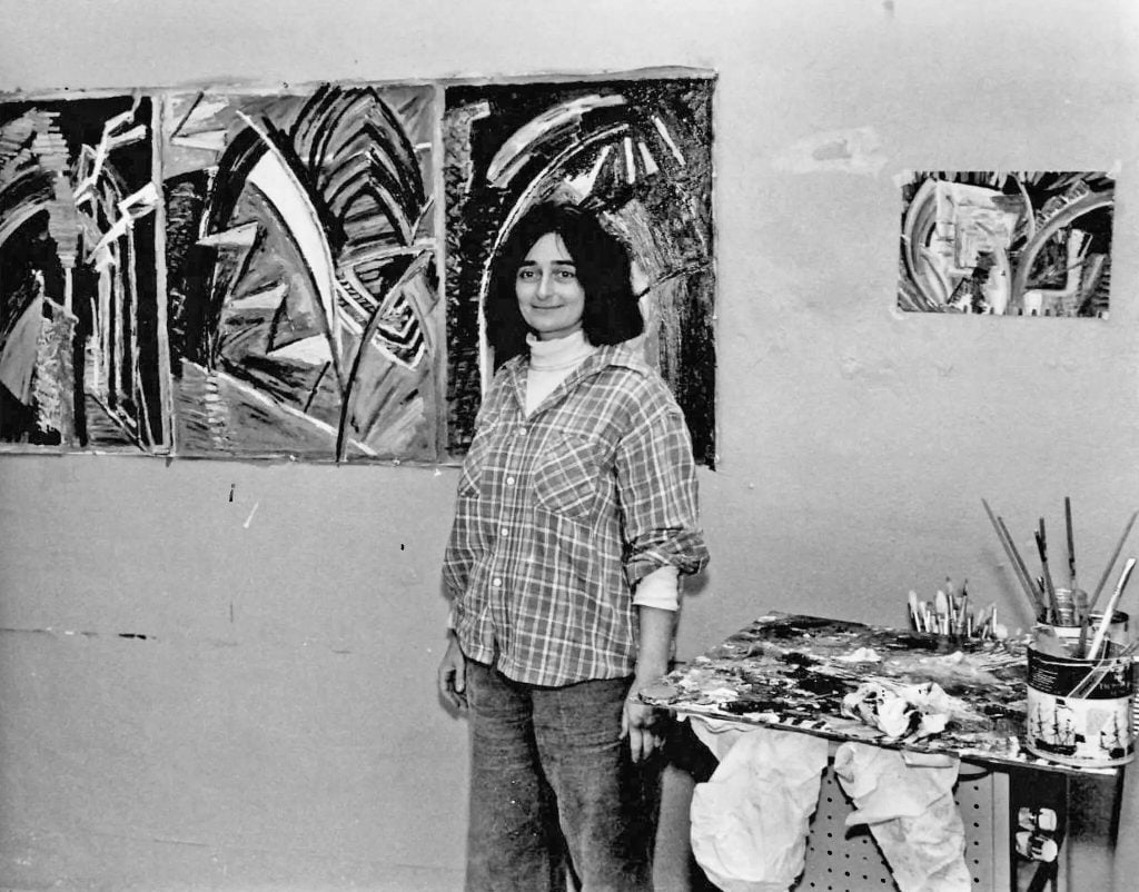 A black and white photo of Katherine Porter in her studio. She is a young white woman with shoulder length dark hair standing front of abstract paintings hanging on the wall and a table piled with brushes and paint. 