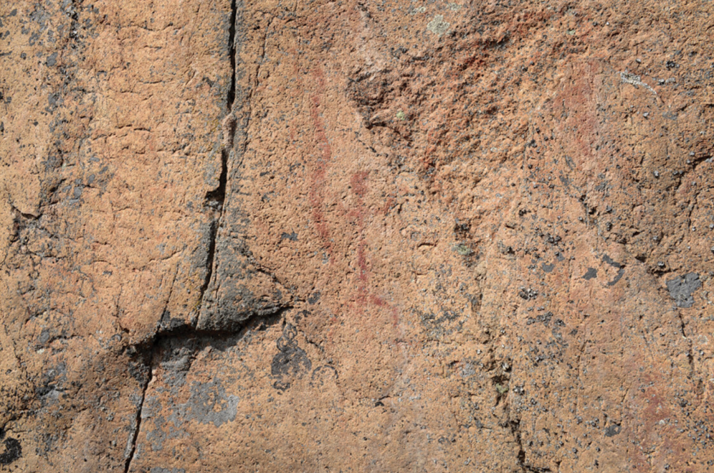 A photograph of a rock facade with a faint red painting of a stick figure person holding a stick figure snake.