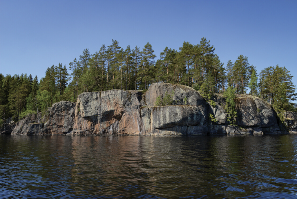 A photograph of a rock formation jutting over a lake with trees atop it and blue skies above.