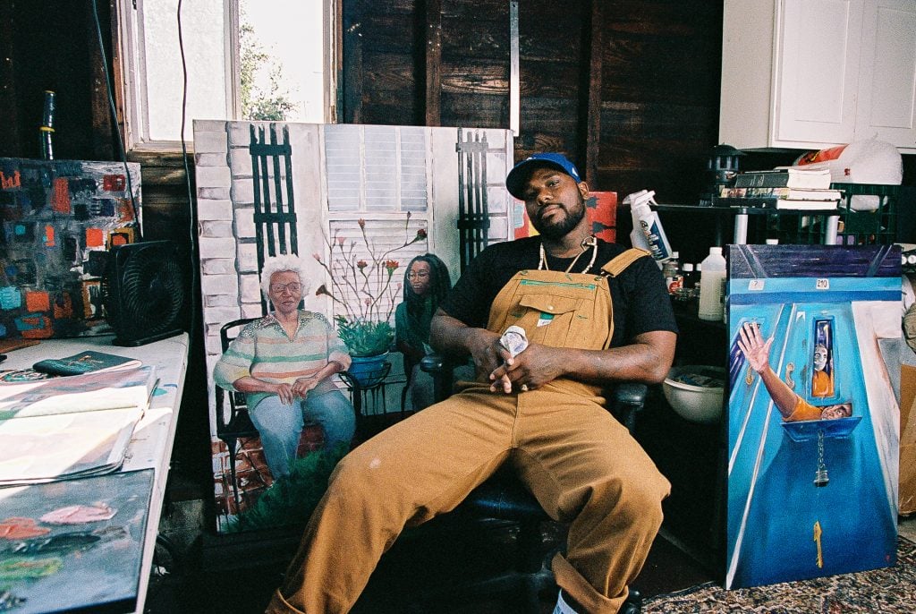 A portrait of a Black artist in his studio with his hands clasped in his lap in front of his face as he is surrounded by paintings.