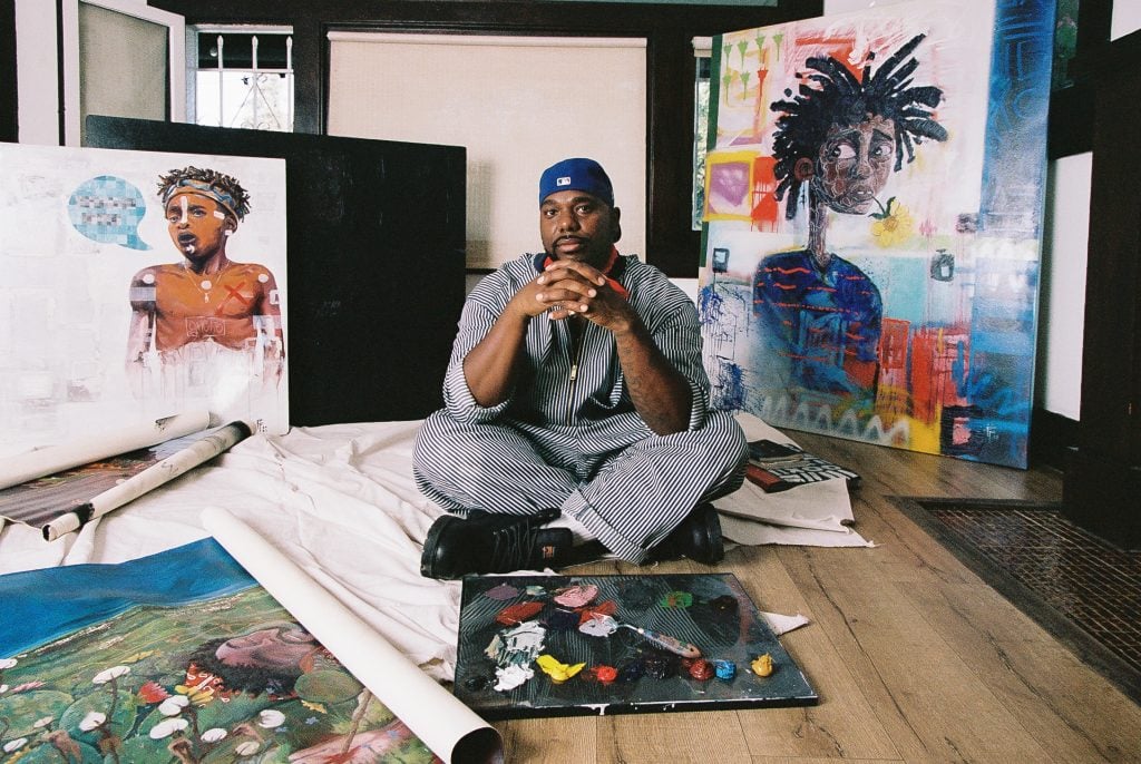 A portrait of a Black artist in his studio with his hands claspes in front of his face as he is surrounded by paintings.