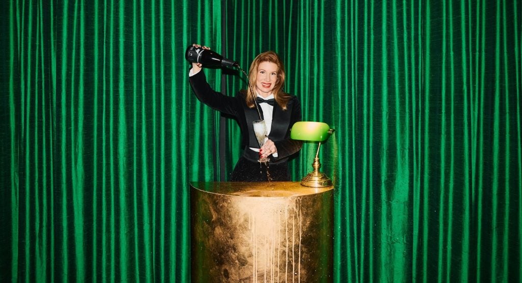 Artist Kirsha Kaechele in a suit pours herself champagne while standing in a lushly appointed space, her Ladies Lounge