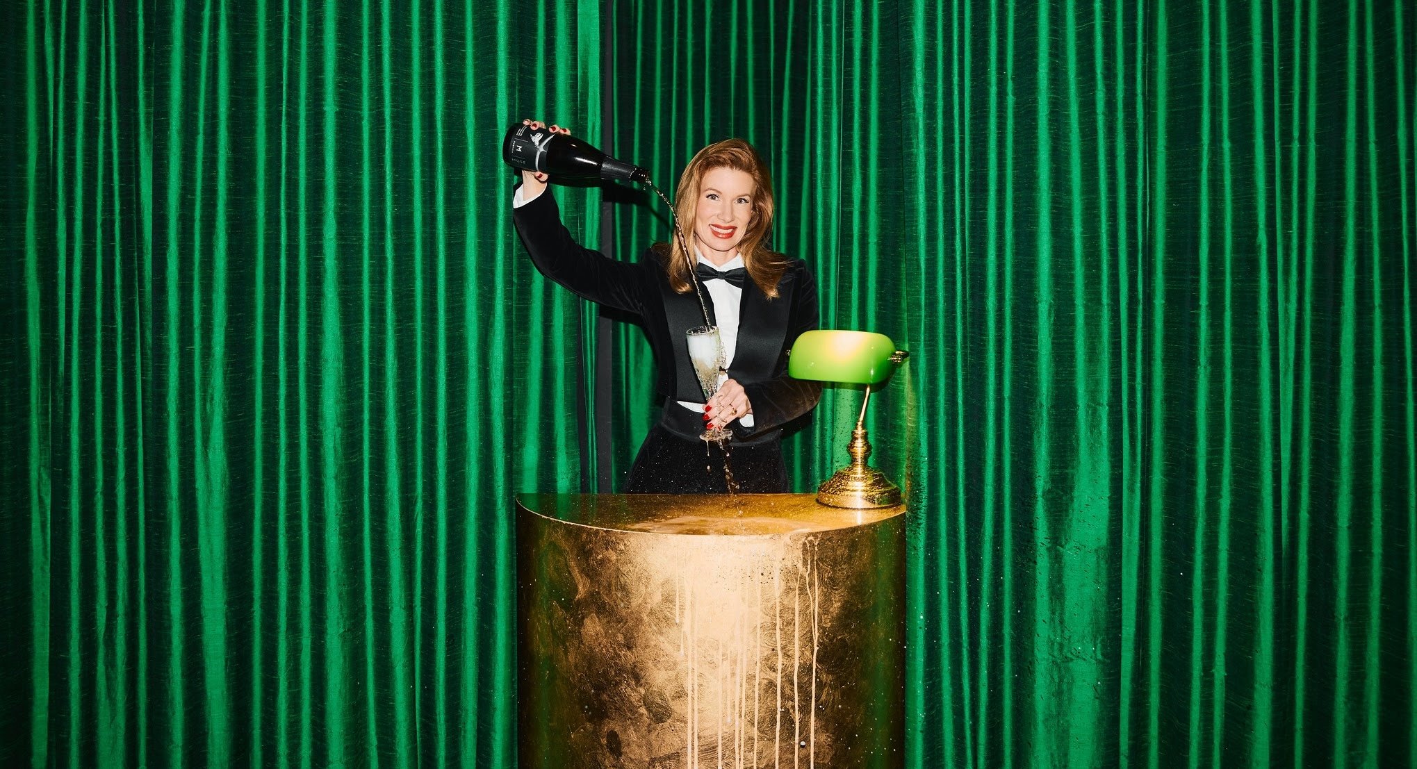 Artist Kirsha Kaechele in a suit pours herself champagne while standing in a lushly appointed space, her Ladies Lounge