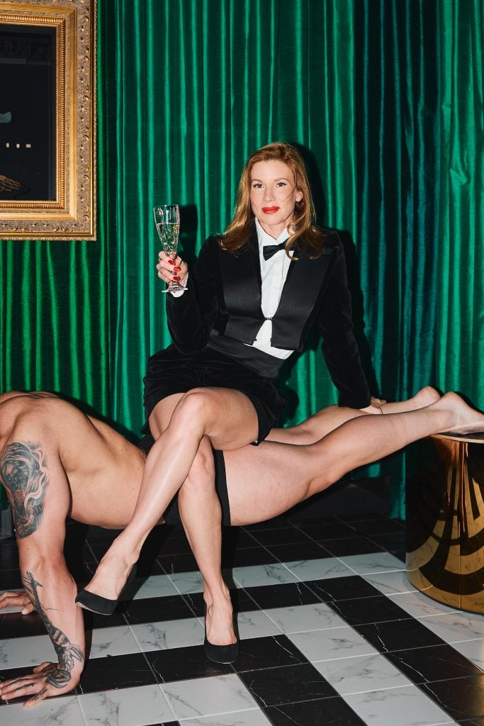 A white woman with a glass of champagne sits on the back of a naked man forming a plank for her to sit on