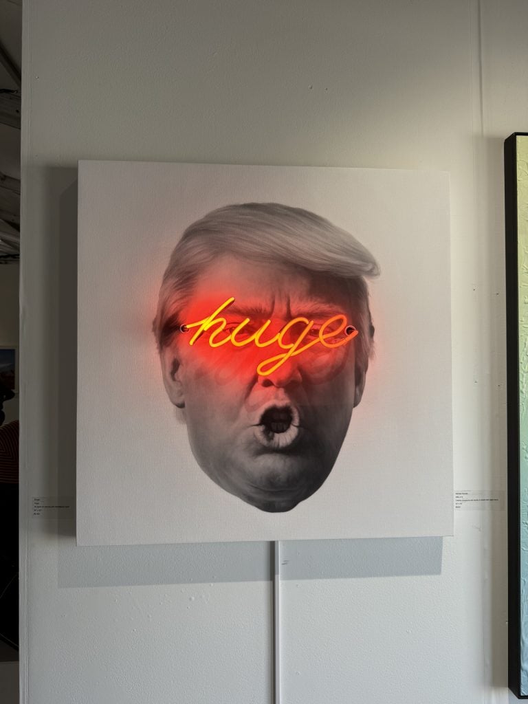 A photograph of an unframed artwork depicting Donald Trump with neon reading "Huge" over his eyes, mounted on a white wall with a cord extending down from it—and one other painting barely visible off to the right.