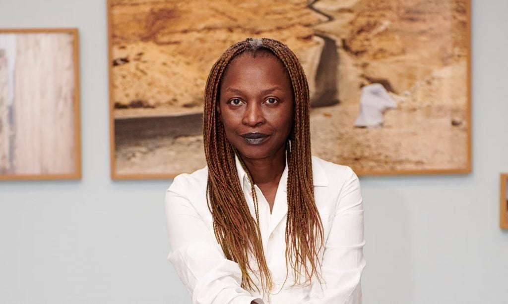 a woman in a white dress with braids sits in front of a wall with artworks